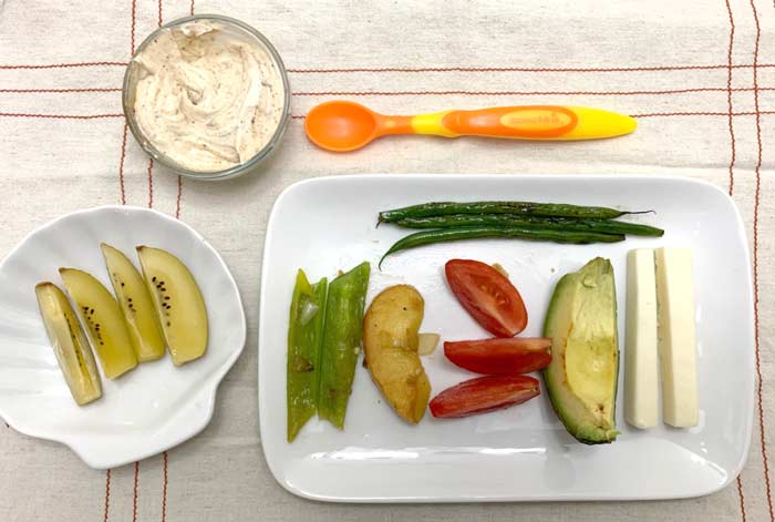 Baby-led weaning meal - Starting top left: a) organic yogurt with peanut butter, b) sliced kiwis, c) sauteed green beans, d) pan-roasted pepper and apple slices, e) garden-fresh tomatoes, f) avocado, g) sheep cheddar. The spoon pictured at the top was one we were gifted; it didn't work well because the handle was too thin. 