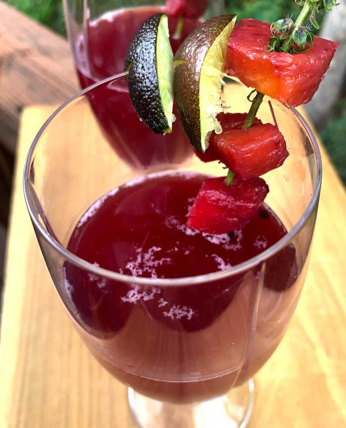 A glass of fermented chicha morada garnished with slices of Australian blood limes. We also used sprigs of purple basil flowers to hold the fermented pineapple and pear pieces. You can also see a few of the fermented coriander seeds floating on the surface of the chicha.